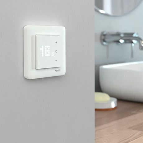 Valkoinen kosketusnäyttötermostaatti harmaalla seinällä, taustalla kromihana ja pesuallas.