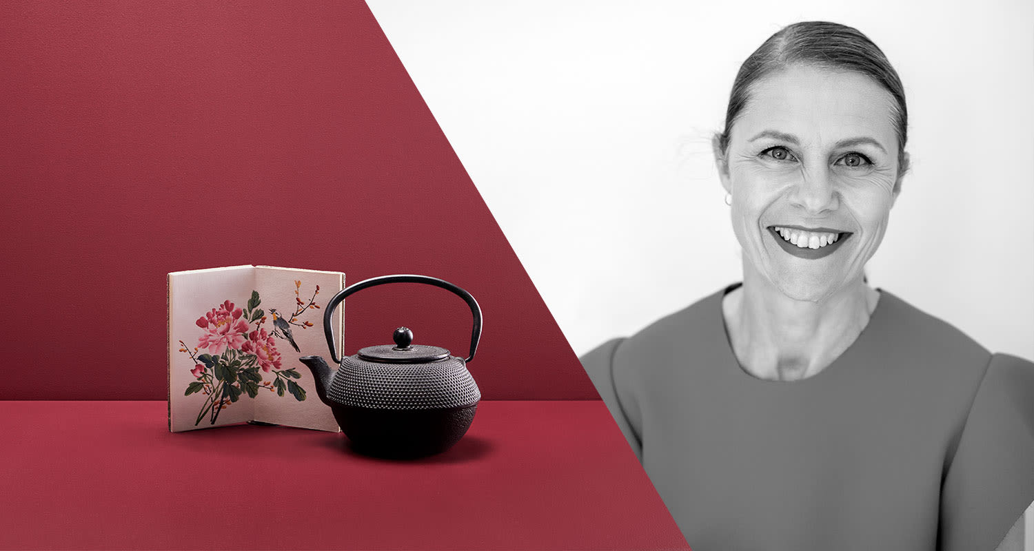 Punaisella alustalla valurautainen teepannu ja kukkakuvioitu näyttöpaneeli; harmaasävyinen puku.