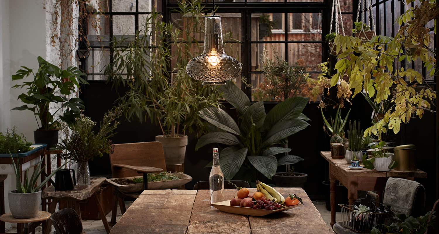 Rustiikkinen sisäpuutarha täynnä viherkasveja, puupöytä hehkulampun ja hedelmävadin kanssa.