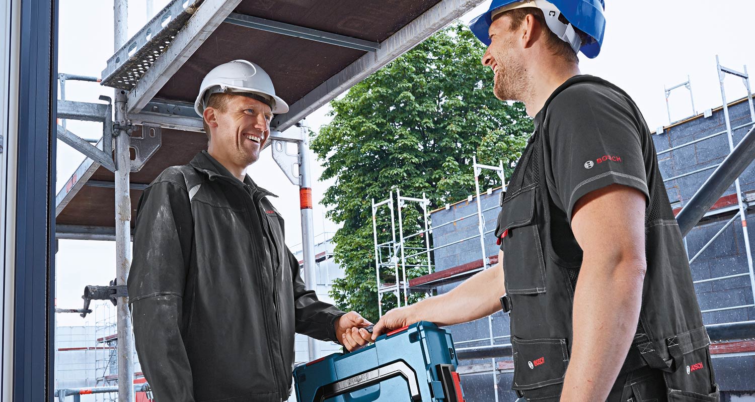 Kaksi kypäräpäistä rakennustyöntekijää kantaa sinistä työkalulootaa rakennustelineiden edustalla.