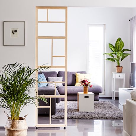 Vaalea olohuone: harmaa sohva värityynyillä, puuseinäke, vihreä palmu ja kukkavaasi.