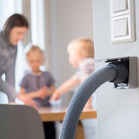 Harmaa keskusimurin letku seinäliitännässä, taustalla äiti ja lapset keittiön pöydän äärellä.