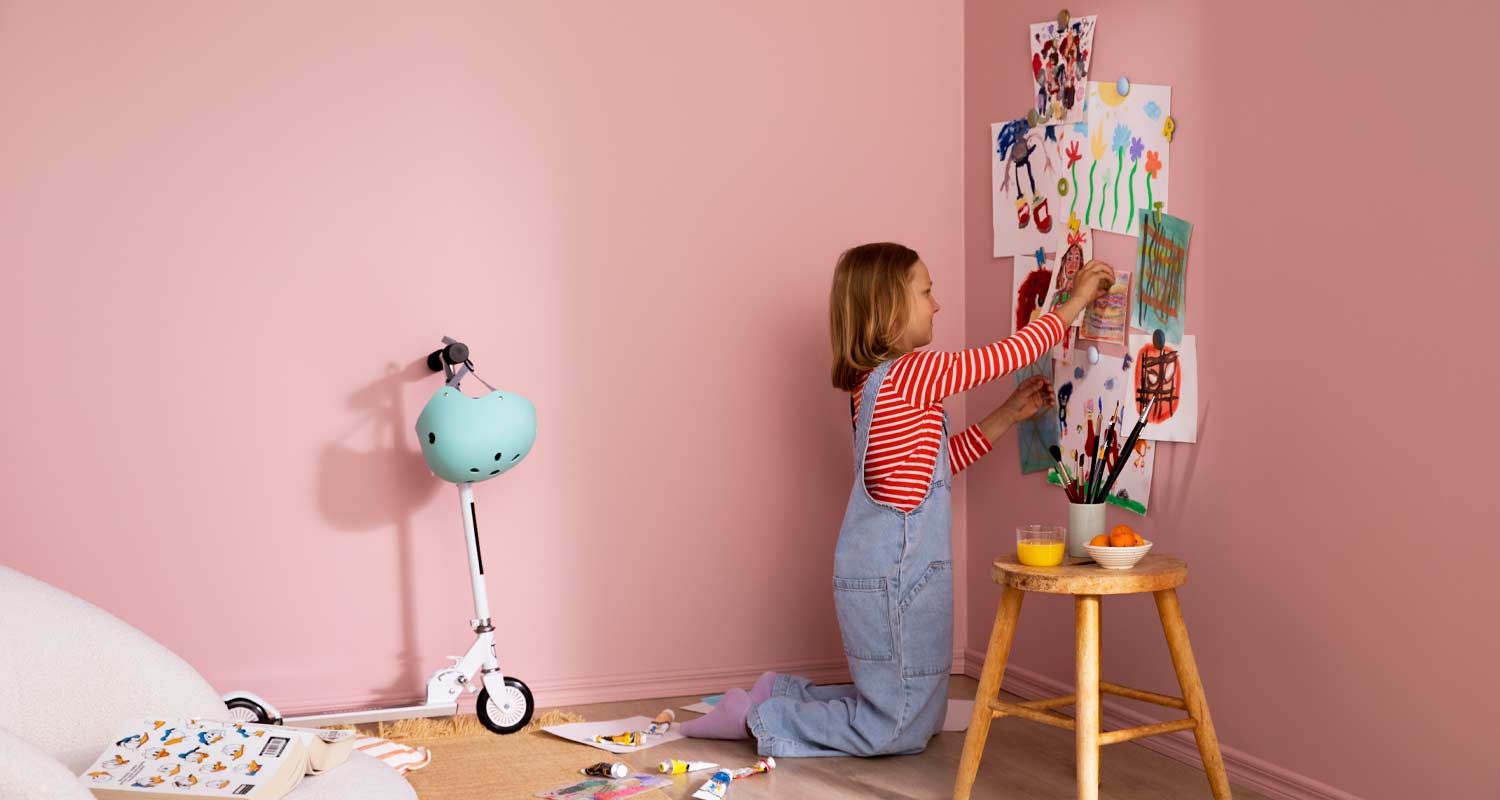 Vaaleanpunaisessa nurkassa lapsi kiinnittää piirustuksia seinälle, potkupyörä, välineet ja välipala