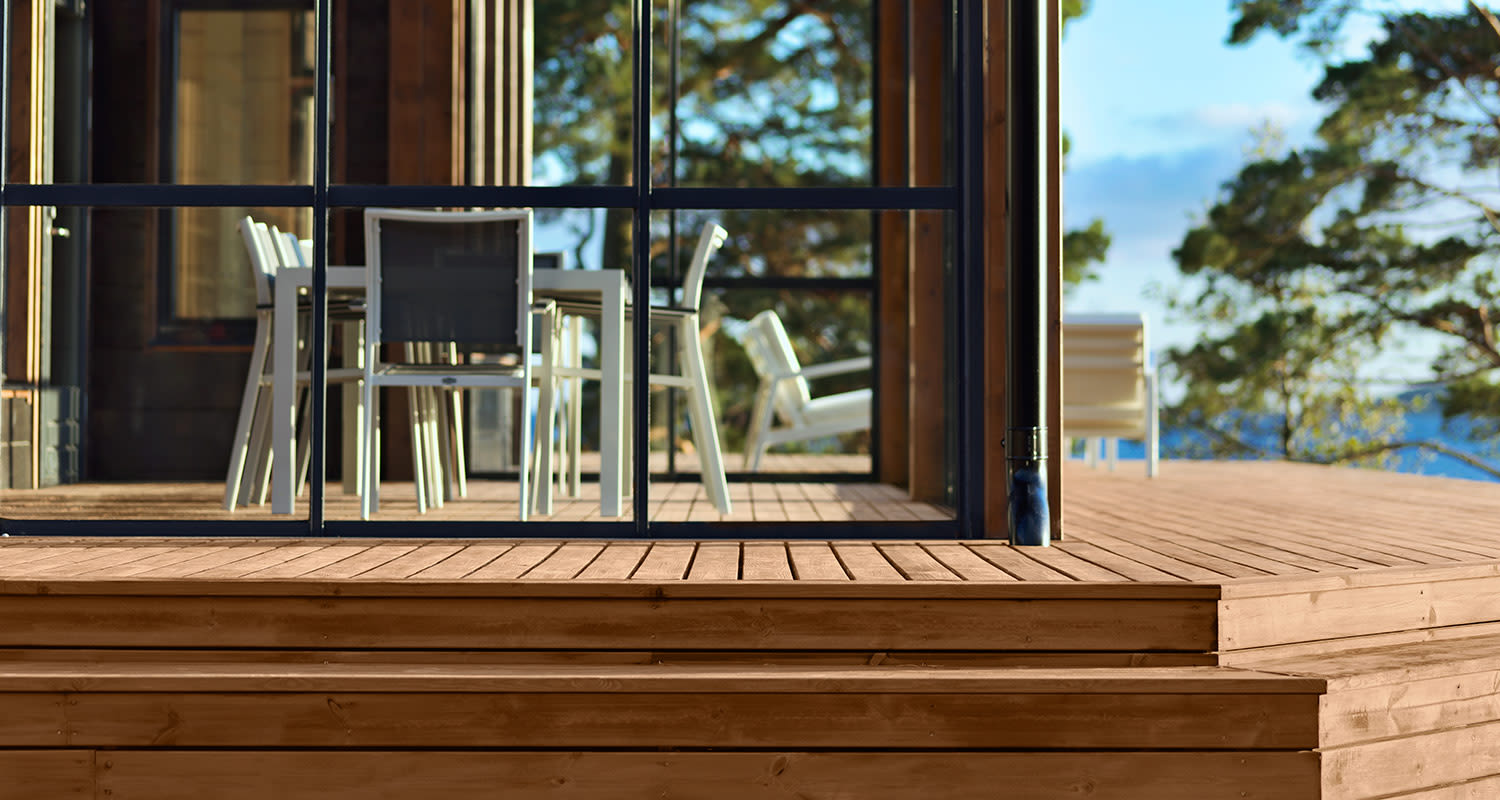 Vaalea puulattia ja lasiseinä, valkoiset terassituolit, metsä- ja järvinäkymä