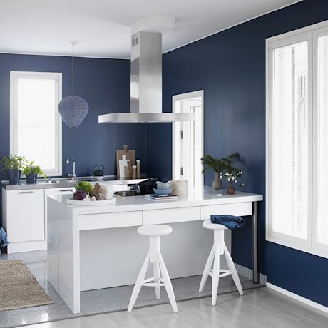 Moderni keittiö tummansinisin seinin, valkoinen saareke, kaksi baarituolia ja liesituuletin.