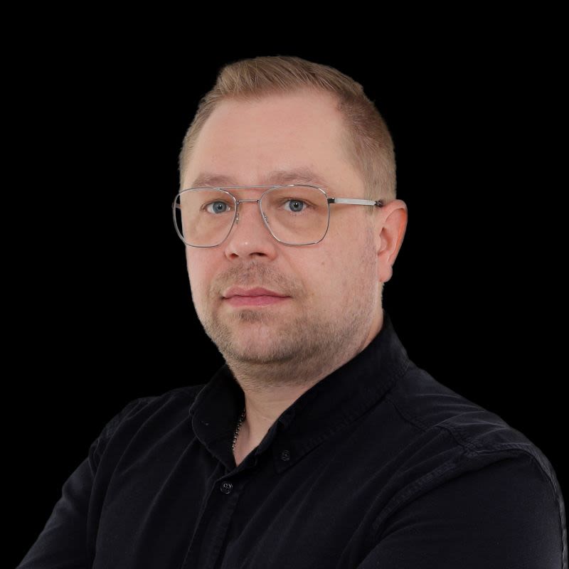 Henkilö lyhyellä vaalealla tukalla ja silmälasikehyksellä mustassa paidassa mustalla taustalla.