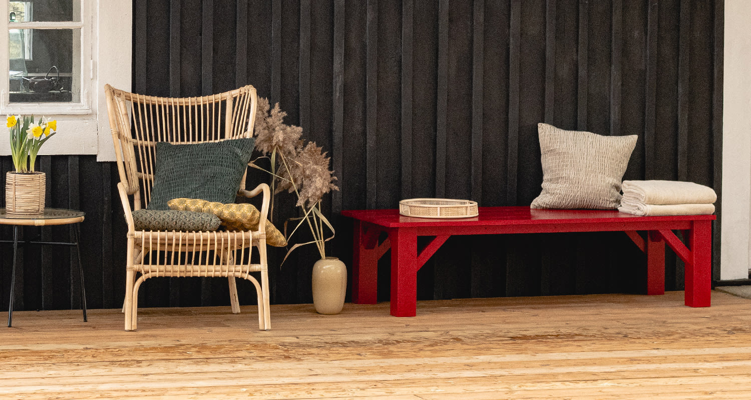 Rottinkinojatuoli vihreä- ja keltatyynyillä, narsissit pöydällä, puna-penkki tyynyine.