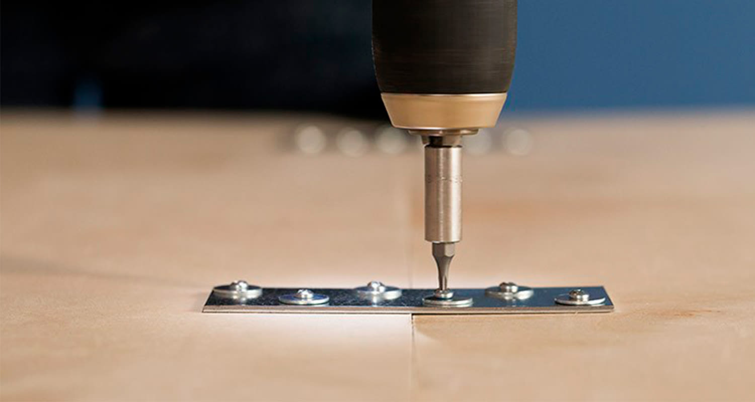 Porakoneen metallinen kärki ruuvaa hopeista liitoslevyä kahden vaalean puulevyn saumassa