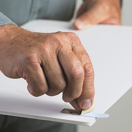 Miehen käsi irrottaa teipin suojakalvoa valkoiselta levypinnalta.