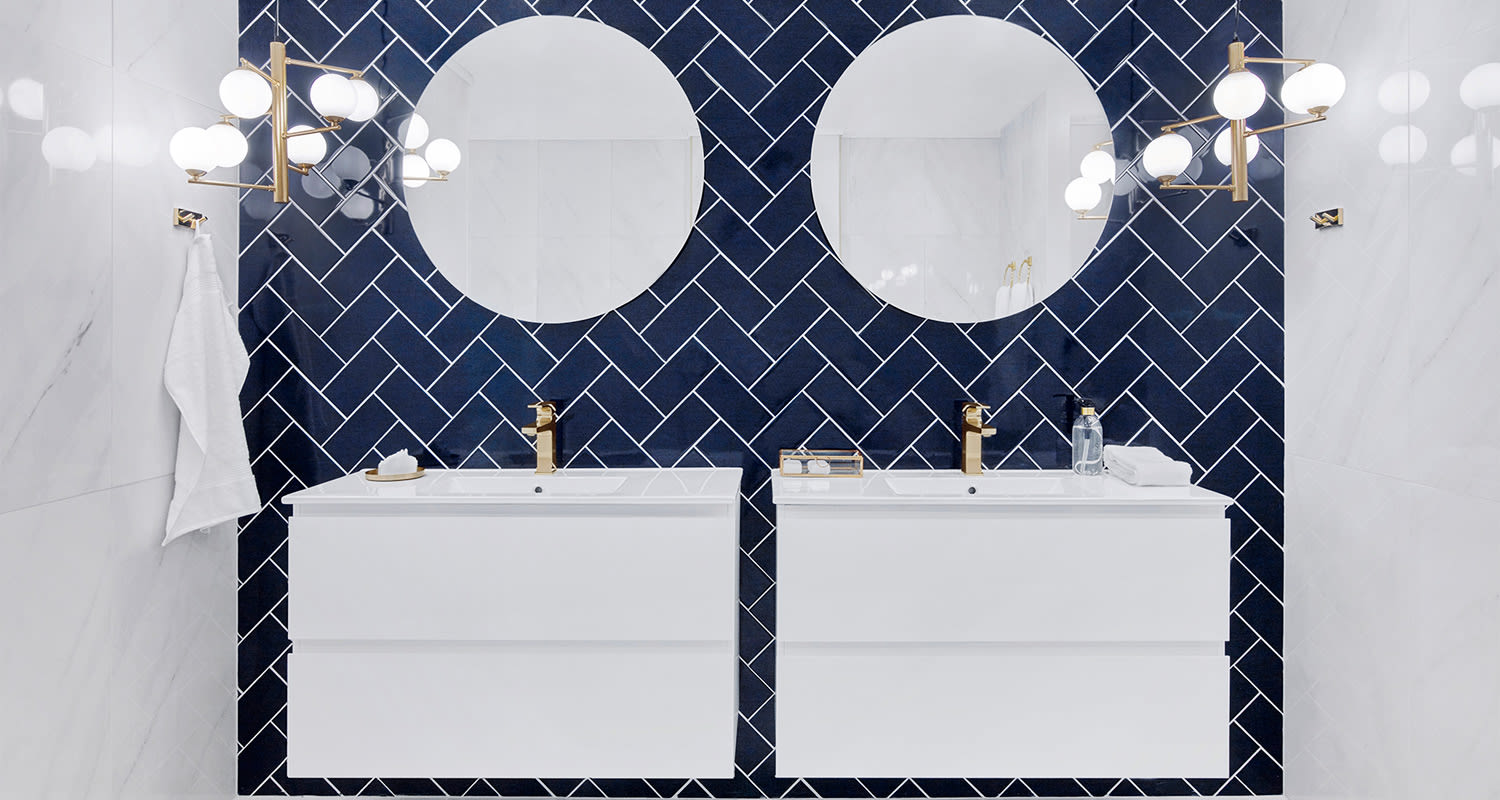 Kylpyhuone, valkoinen allaskaappi, tummansininen kalanruotolaatta, pyöreät peilit kultaiset hanat