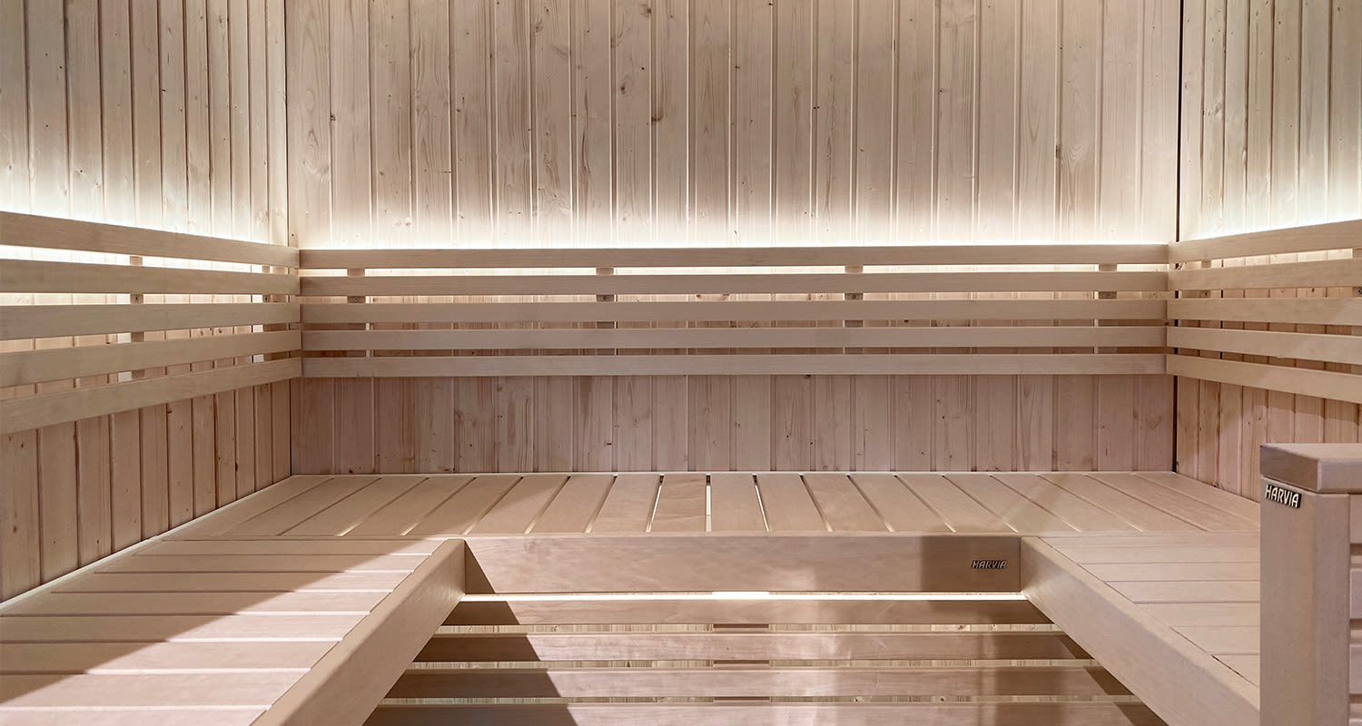 Tyhjä vaalea puusauna, L-muotoiset penkit ja himmeä LED-valaistus seinän takana