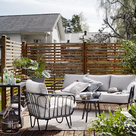 Harmaa ulkosohva, mustakehysnojatuoli ja pikkupöytä puuaidatulla terassilla, viherkasveja.