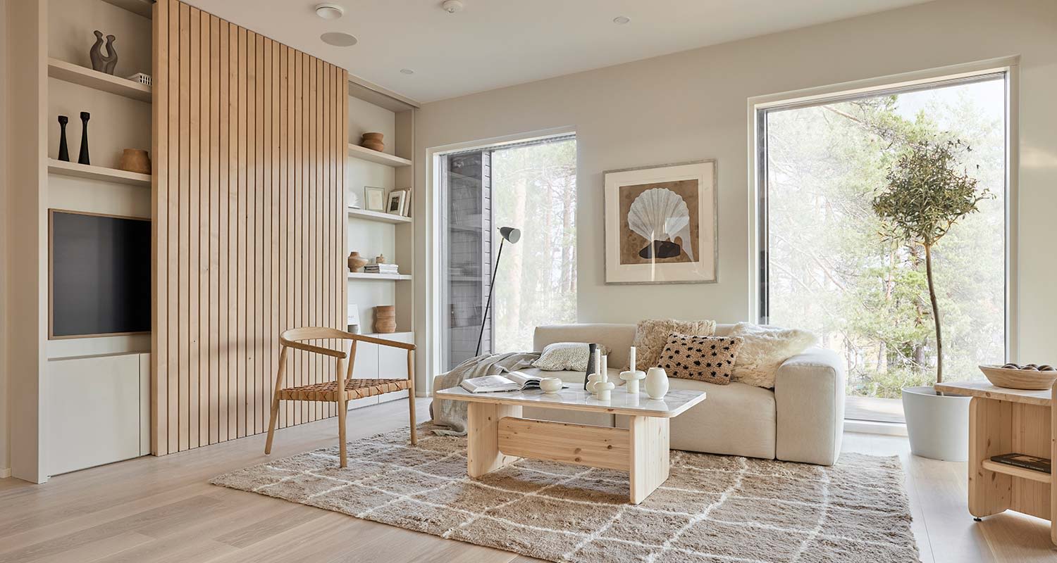 Beige olohuone, puinen sohvapöytä, rottinkituoli ja isot ikkunat metsänäkymällä.