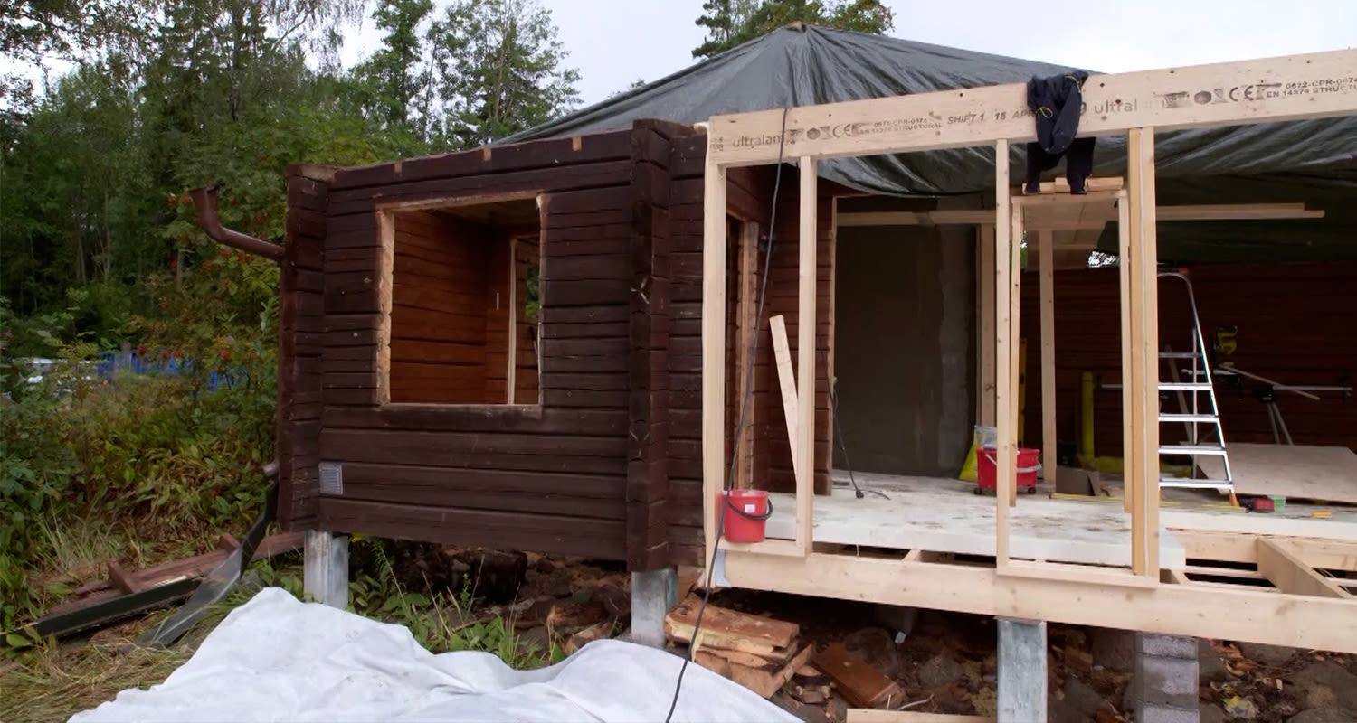 Ruskea hirsimökki metsässä, osa purettuna, sisällä vaalea puurunko, työkaluja ja tikkaat.