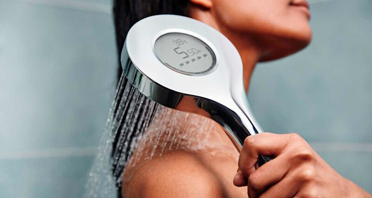 Krominen diginäytöllinen suihkupää suihkuttaa vettä naisen olkapäälle harmaata taustaa vasten.