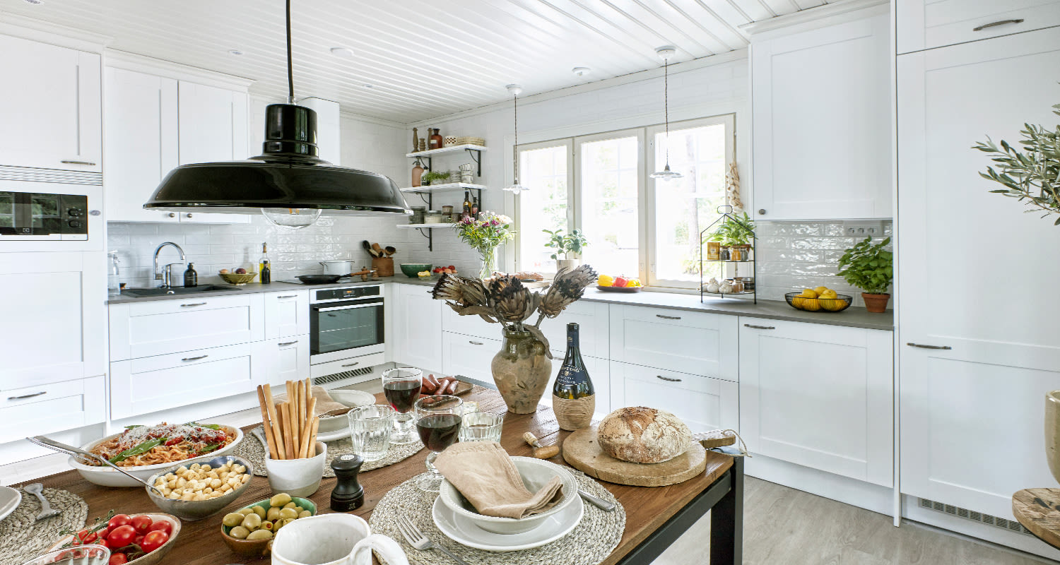 Vaalea maalaiskeittiö mustalla kattolampulla, ruokapöydällä leipää, pastaa, oliiveja ja viiniä.