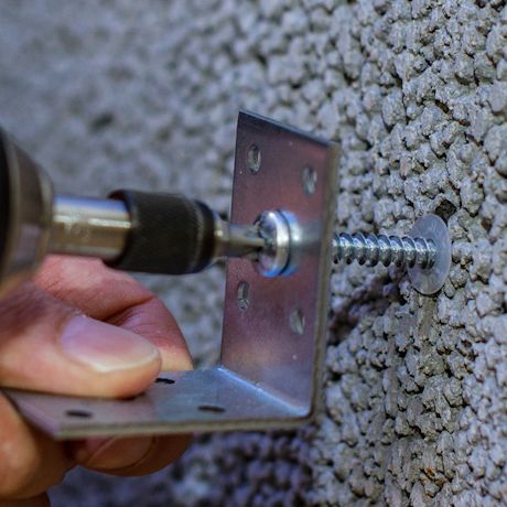 Kädessä pidettävä hopeinen kulmatuki ruuvataan karkeaan harmaaseen seinään