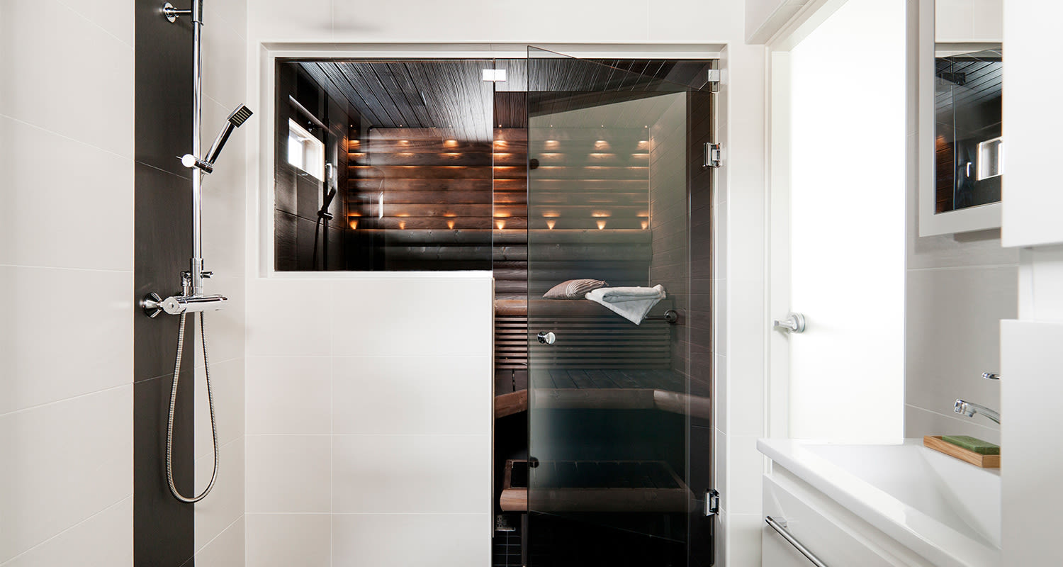 Valkoisessa kylpyhuoneessa krominen suihku ja lasiovellinen, valaistu tumma puusauna.