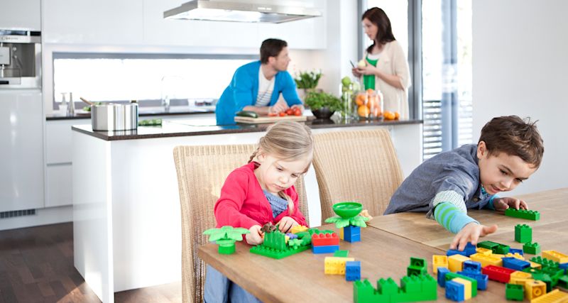 Lapset leikkivät värikkäillä Duplo-palikoilla puupöydän ääressä valkoisessa keittiössä.