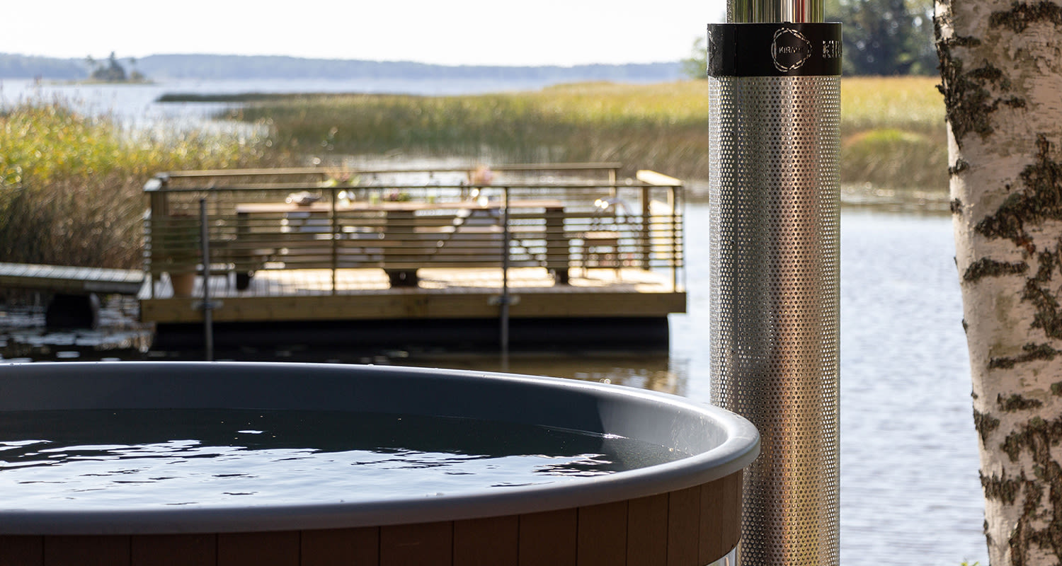 Harmaareunainen puulämmitteinen palju järvenrannalla, metallinen piippu ja puulaituri.