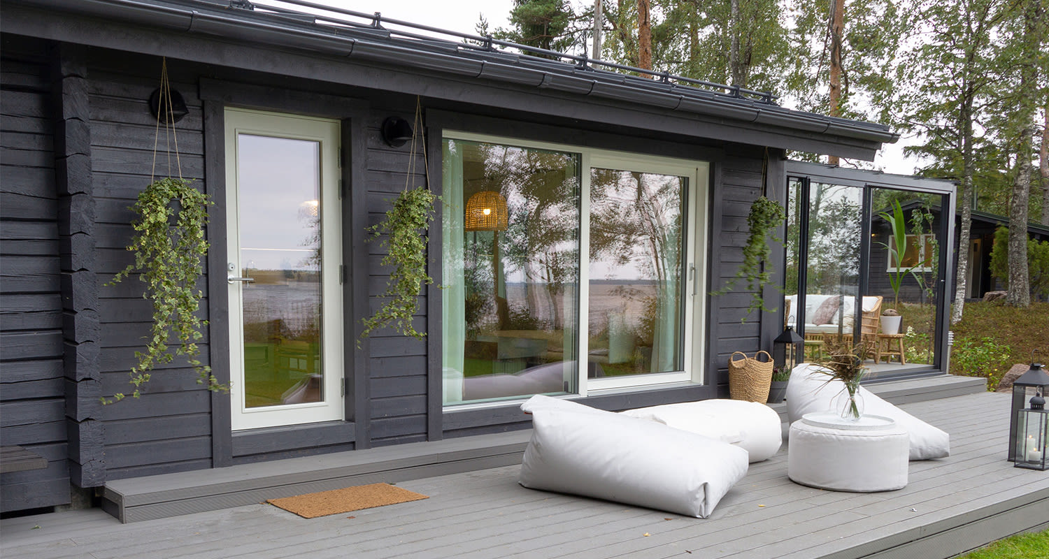 Tummanharmaa puutalo lasiovilla, harmaa terassi valkoisilla tyynyillä, roikkuvat viherkasvit.