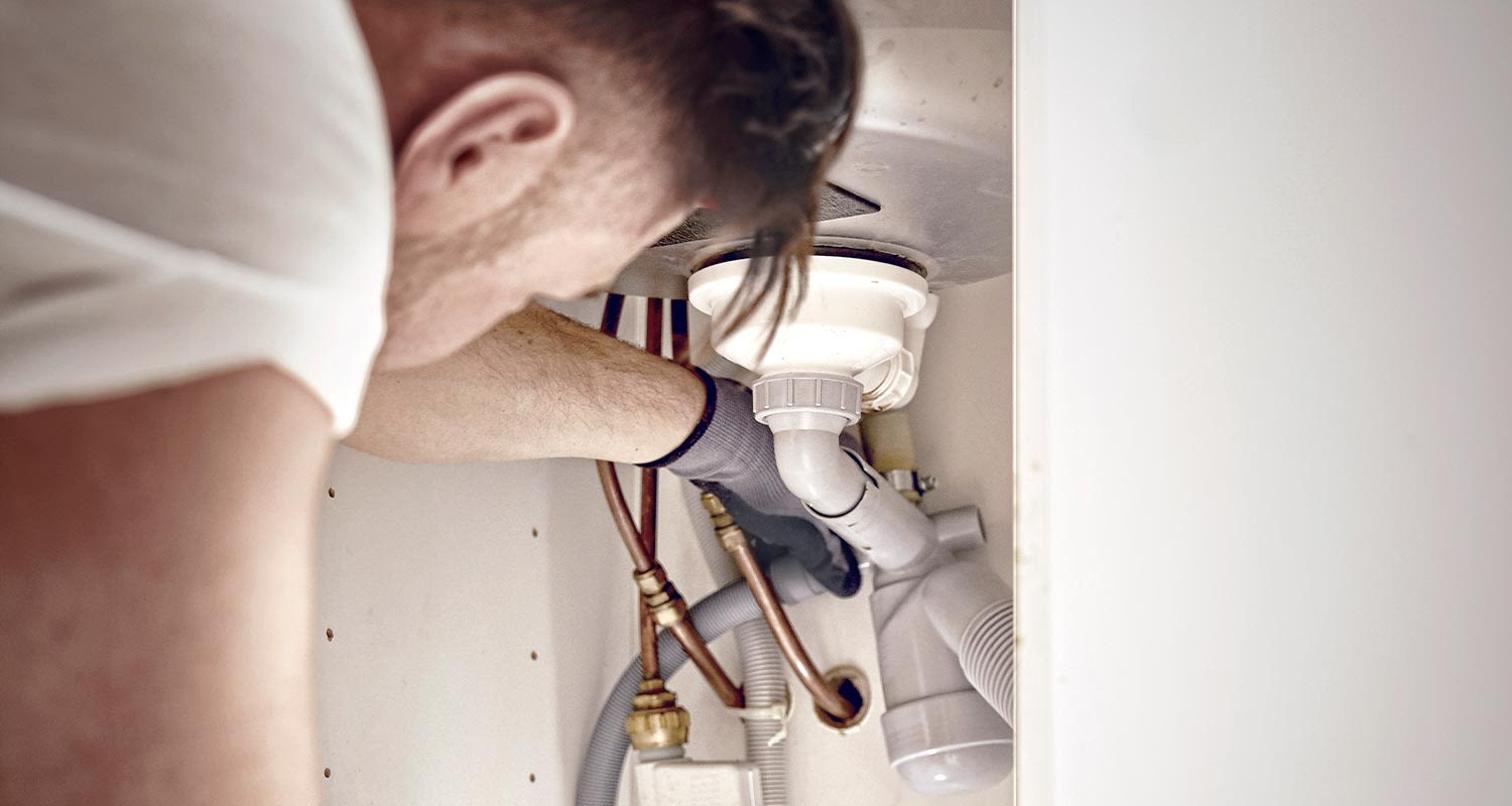 Mies korjaa tiskialtaan alla kupari- ja harmaita muoviputkia mustissa käsineissä.