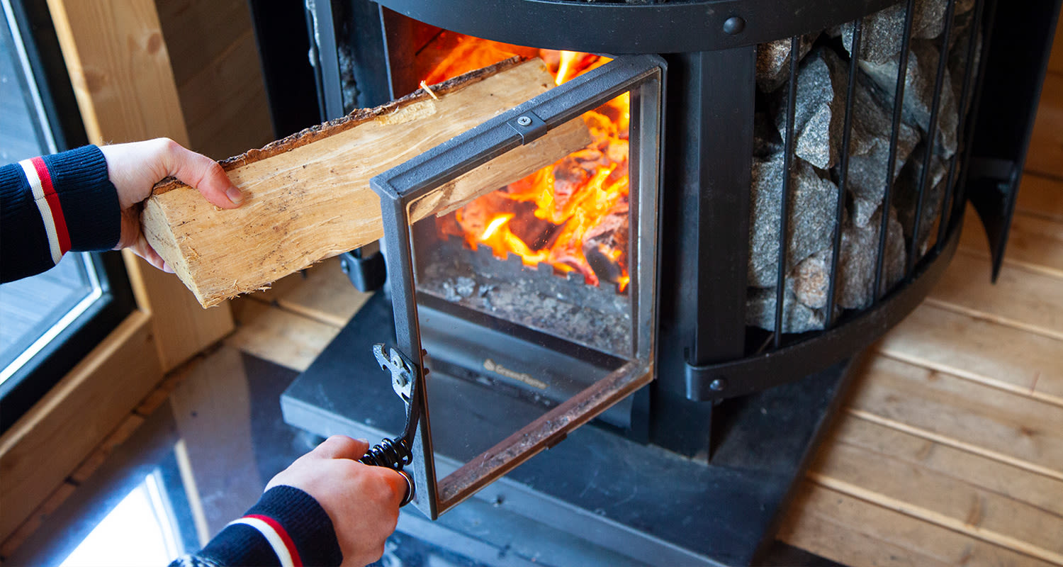 Kädet työntävät halkoa saunan kiukaan tulipesään, näkyvissä oranssit liekit ja kiukaan kivet.
