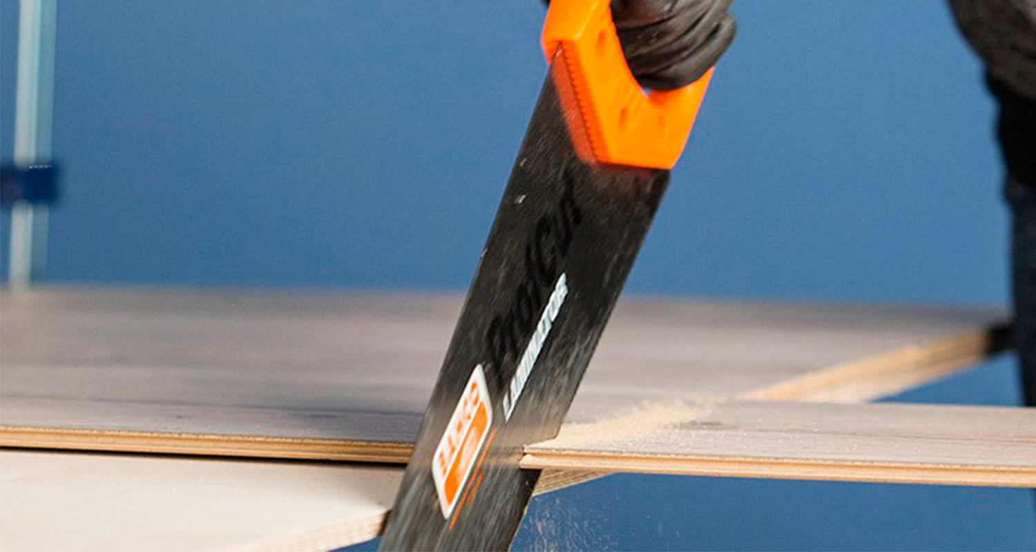 Mustahansikkainen käsi sahaa oranssikahvaisella käsisahalla vanerilevyä sinistä taustaa vasten.