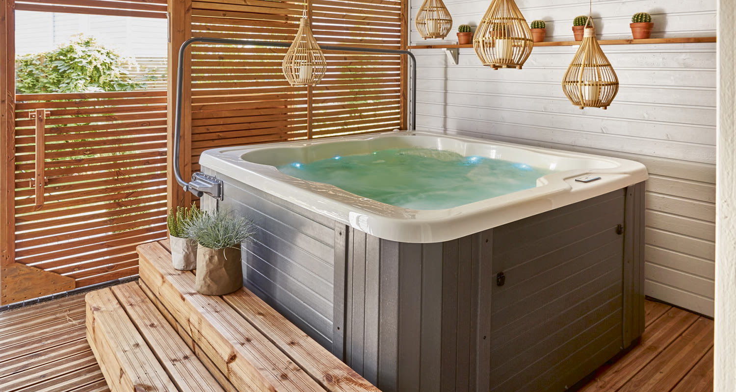 Harmaa kylpytynnyri sinisellä vedellä puisella terassilla rottinkivalaisimien ja kasvien kera