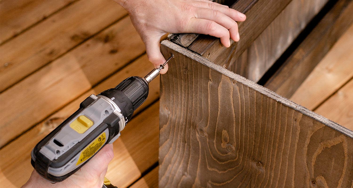 Kädet ruuvaavat harmaamustalla akkuporakoneella tummanruskeaa puulautaa vaalealla puulattialla.