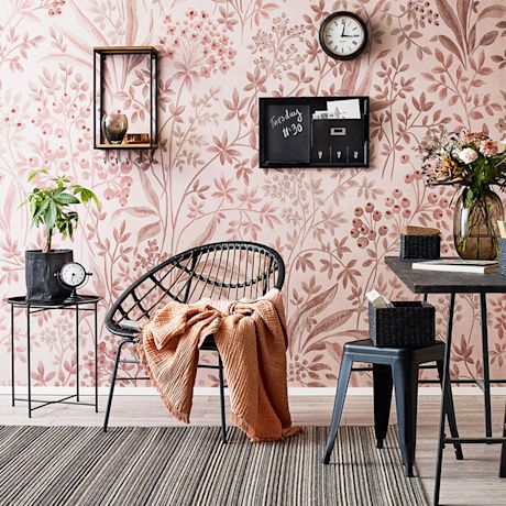 Roosa kukkakuvioinen tapetti, musta rottinkituoli persikkaviltillä, pieni pöytä ja kello