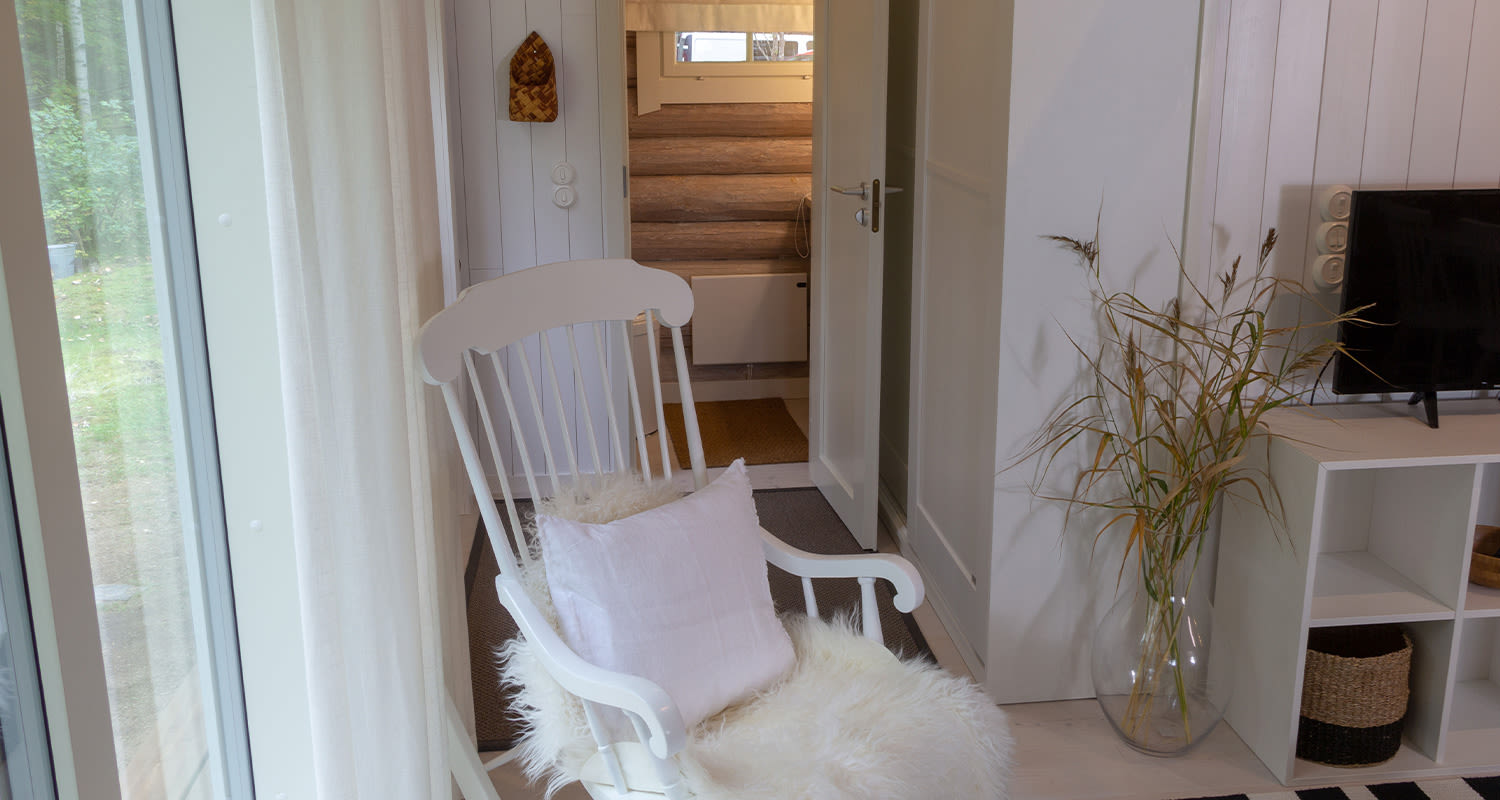 Lasioven vieressä valkoinen keinutuoli lampaankarvalla ja roosatyynyllä, maljakossa kuivaruokoja