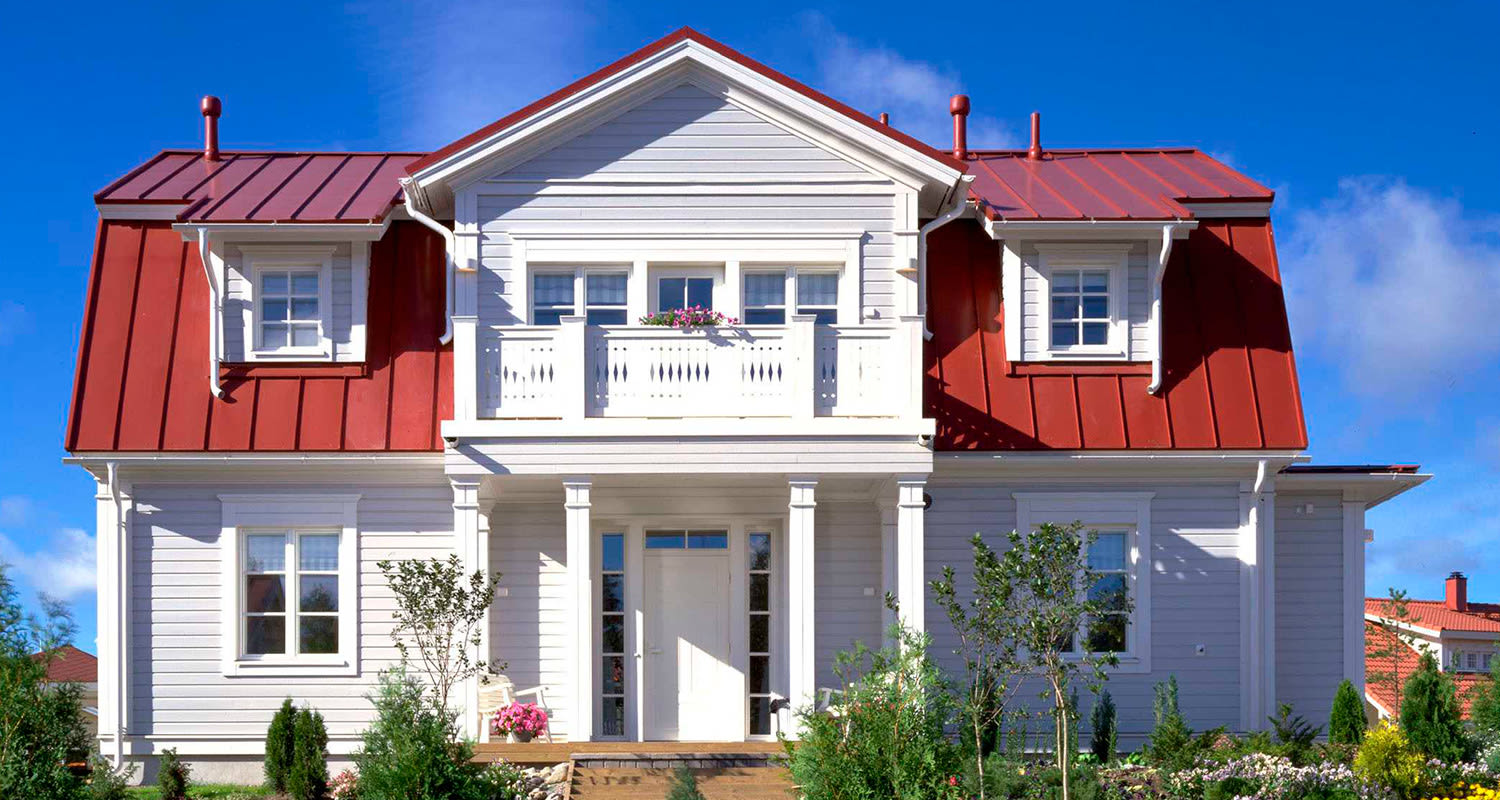 Valkoinen puutalo punaisella peltikatolla, parvekkeella ja kukkivin pensain reunustetulla pihalla