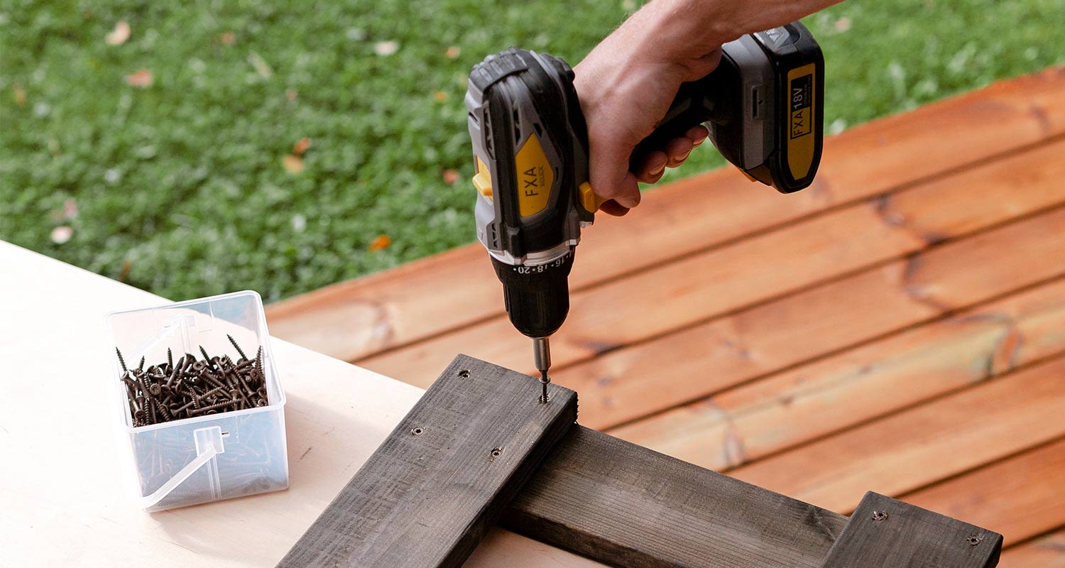 Keltamustalla akkuporakoneella ruuvataan mustaa puupalaa, vieressä muovirasia ruuveja