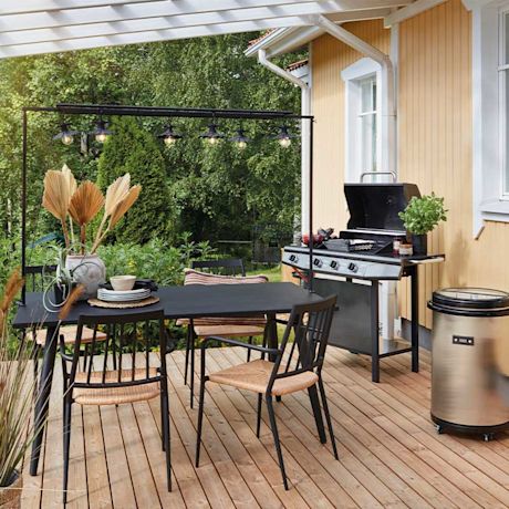 Vaaleankeltaisen puutalon edessä terassi: musta pöytä, punoksiset tuolit, grill ja vihreä puutarha.