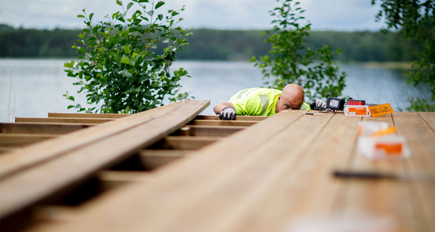 Alt: Rakennustyömies neonkeltaisessa paidassa asentaa puisia lankkuja järven rannalla.