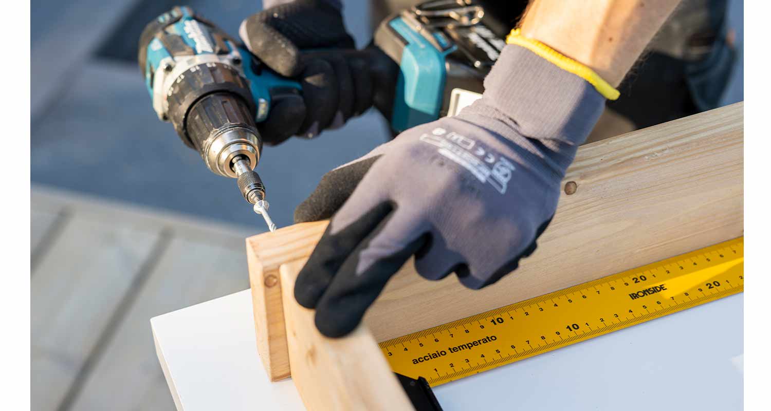 Musta-hansikkainen käsi poraa puupalikkaa sinisellä porakoneella keltaisen viivaimen vieressä