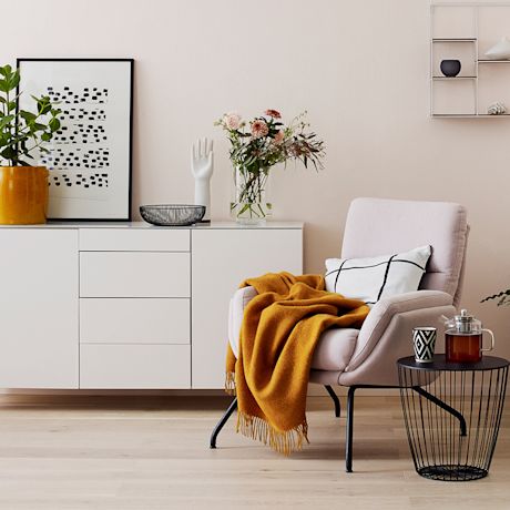 Vaaleanpunaisen seinän edessä vaalea nojatuoli keltaisella viltillä ja musta sivupöytä teekannulla.