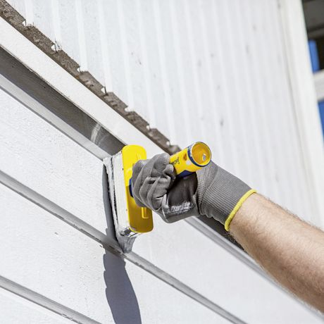 Käsineellinen käsi silottaa keltaisella lastalla harmaata saumamassaa valkoisessa puuverhossa