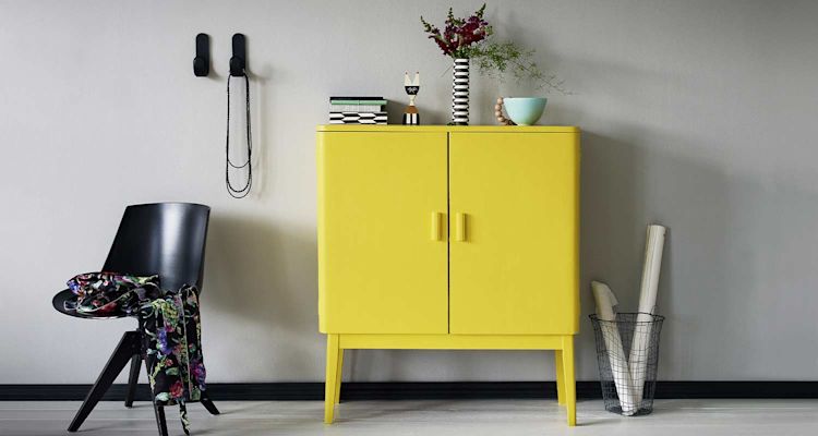 Keltainen lipasto harmaata seinää vasten, musta tuoli kukkahuivilla, koristeita ja rullia korissa.