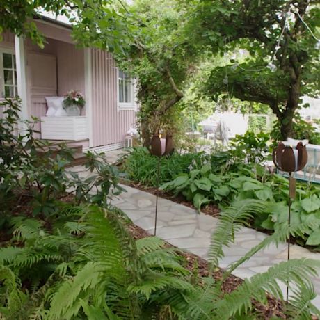 Kivipolku kaartuu rehevän vihreän puutarhan halki kohti vaaleanpunaista mökkiä.