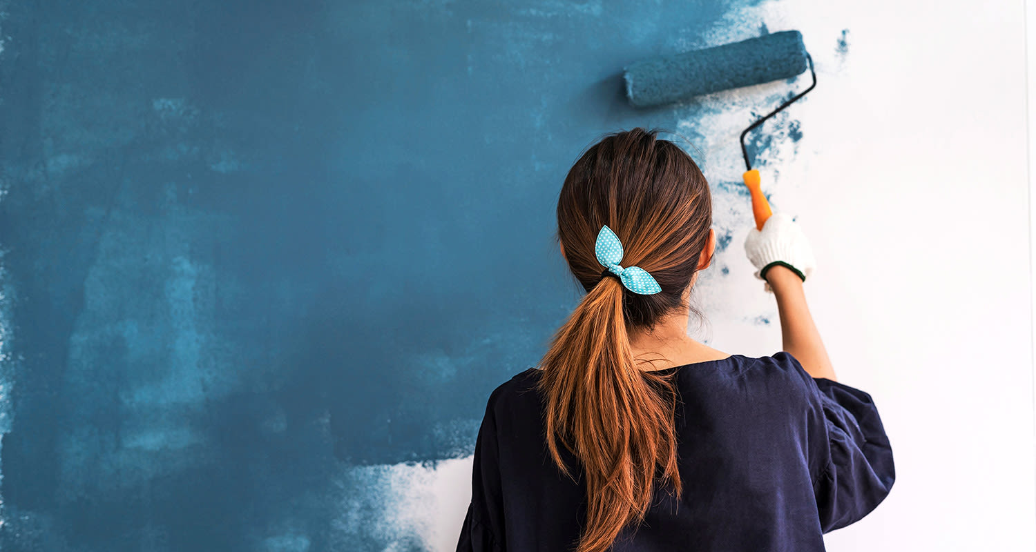 Nainen selin maalaa valkoista seinää tummansinisellä maalirullalla, turkoosipannalla.