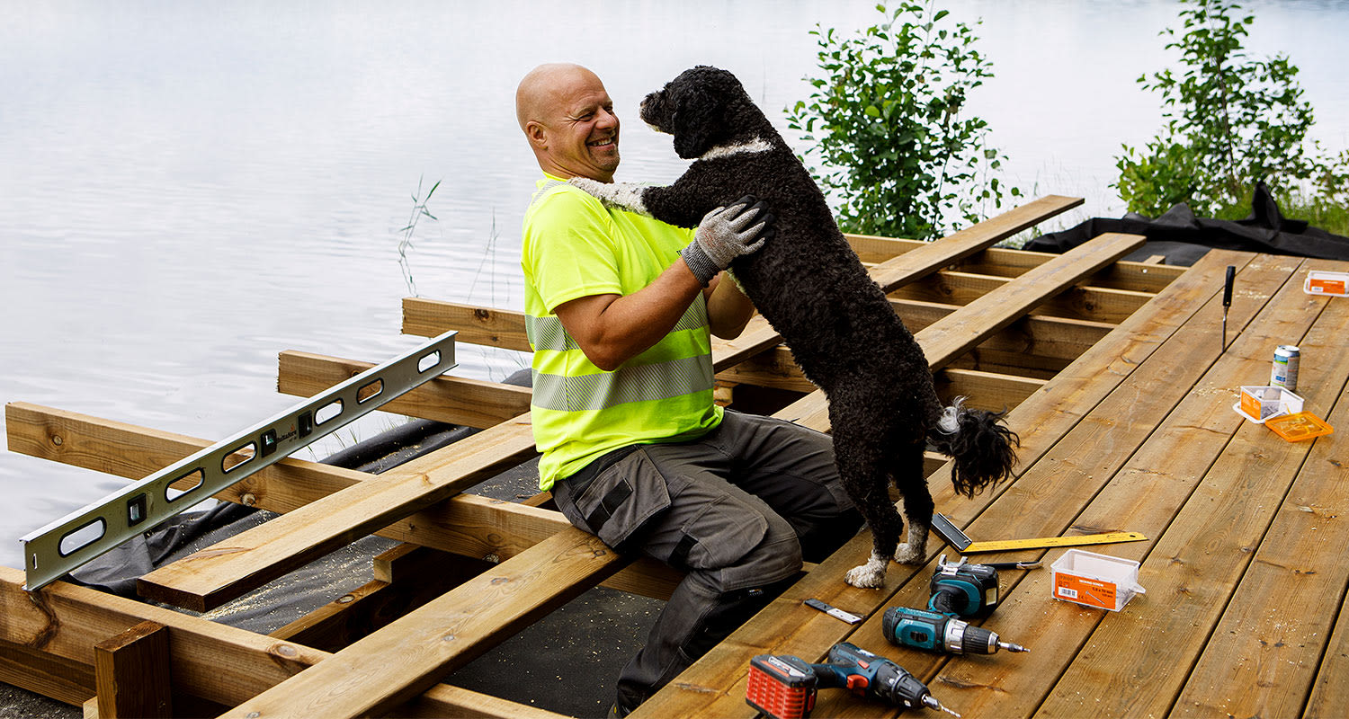 Järven rannalla puisella laiturilla heijastinpaidassa rakentaja leikkii mustavalkoisen koiran kanssa.