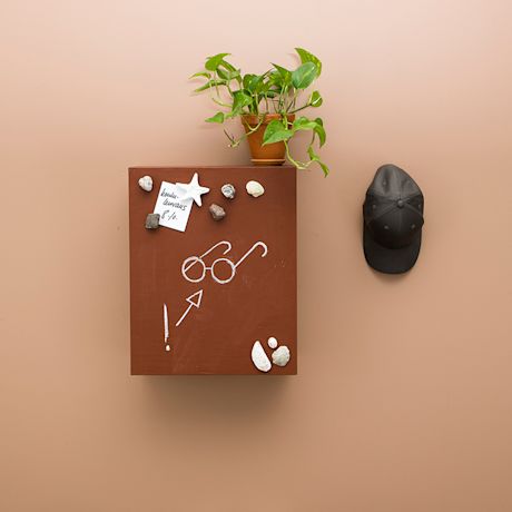 Beigen seinän kuutiohylly liitupiirroksin ja kivimagneetein, päällä viherkasvi, vieressä lippalakki.