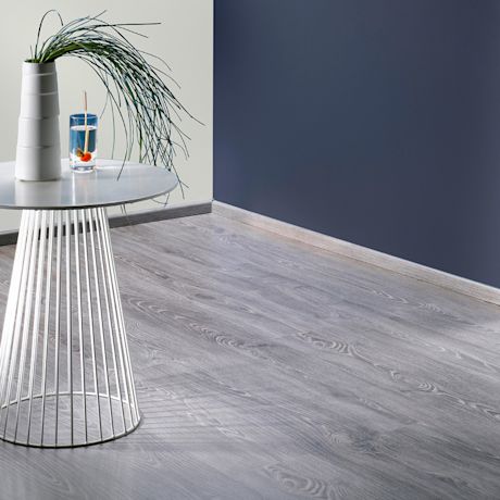 Valkoinen pyöreä sivupöytä harmaalla laminaattilattialla, maljakko ruoholla ja lasi kirsikkavettä.