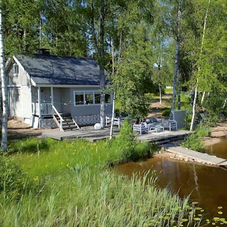 Harmaa kesämökki järven rannalla koivumetsän ympäröimänä, terassi ja laituri veden äärellä.