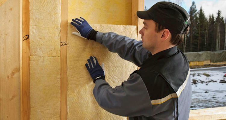 Työmies kiinnittää keltaista lämmöneristettä puurunkoon lumisessa rakennusympäristössä.