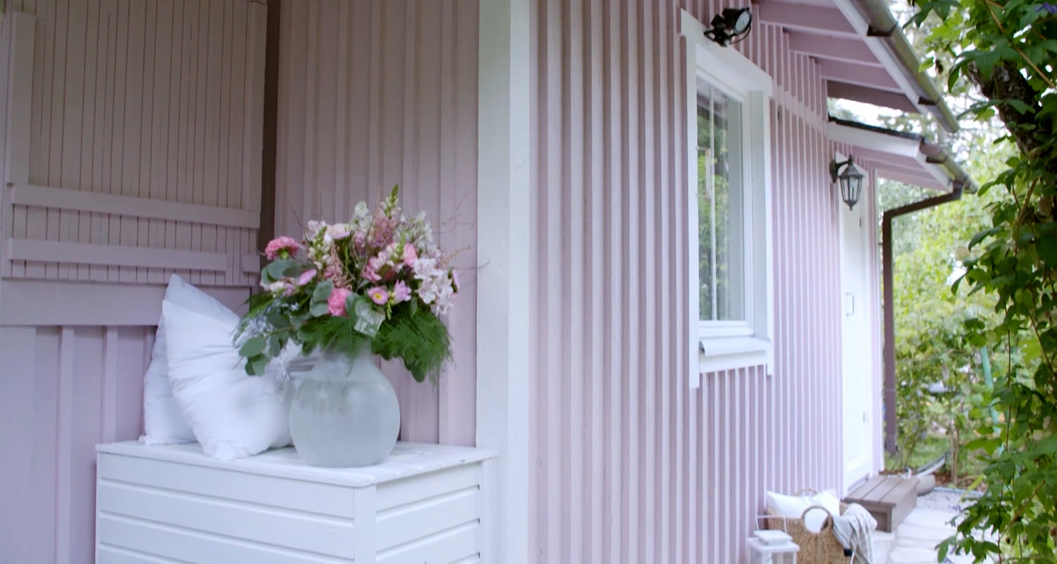 Vaaleanpunaisen puutalon kuistilla valkoinen penkki, tyynyt ja maljakko pinkkejä kukkia