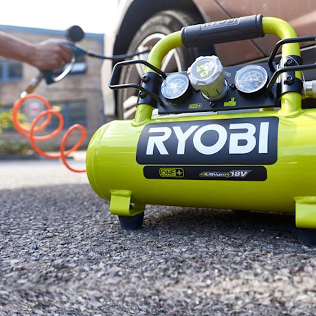 Vihreä Ryobi One+ 18V ilmakompressori mittareilla ja oranssilla letkulla auton renkaassa.