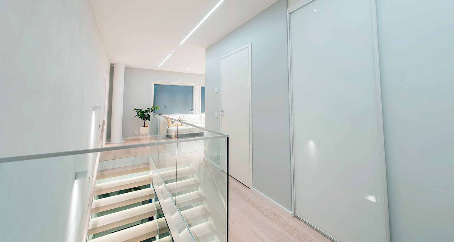 Moderni valkoinen käytävä lasikaiteella, valaistut puuportaat, sohva ja viherkasvi.